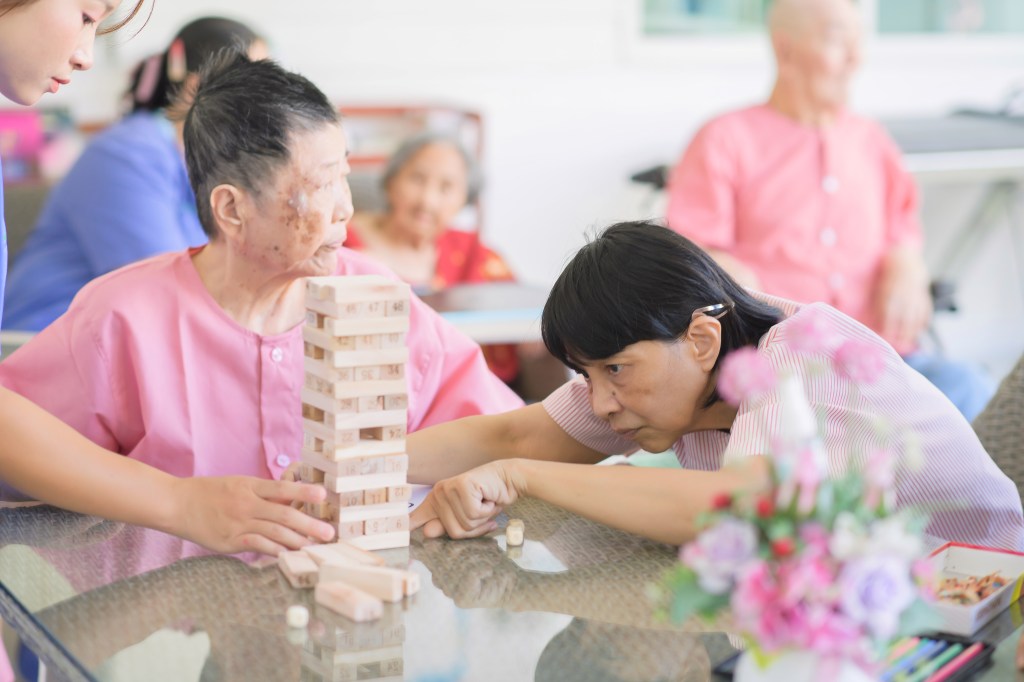 Group of seniors in pink uniforms playing Jenga at a table in a care home.