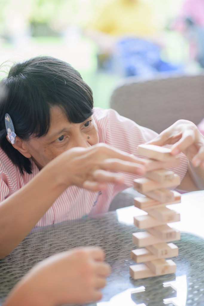 Older woman concentrates as she stacks wooden blocks in a tabletop Jenga-style game outdoors.