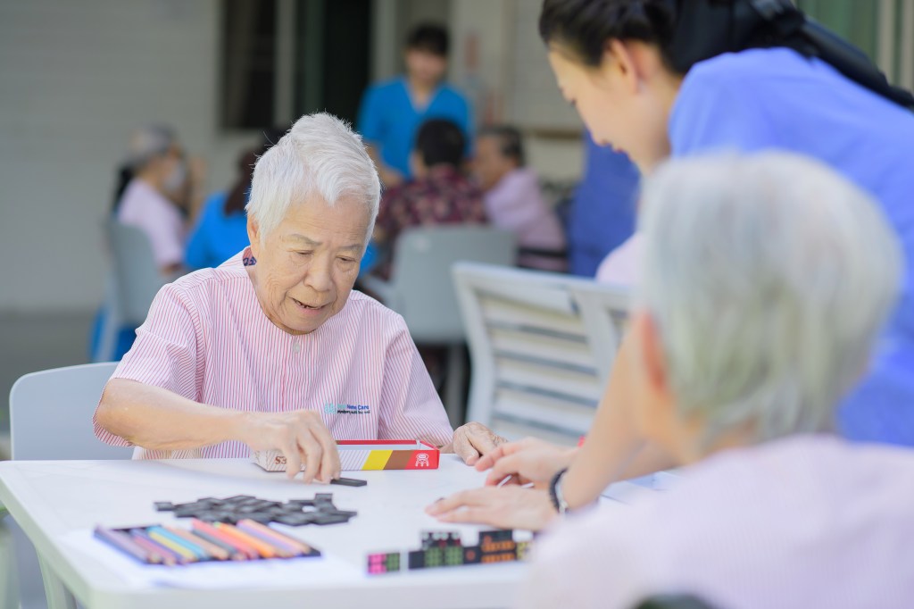 An elderly man concentrates as he arranges small black tiles on a white table during a group activity.